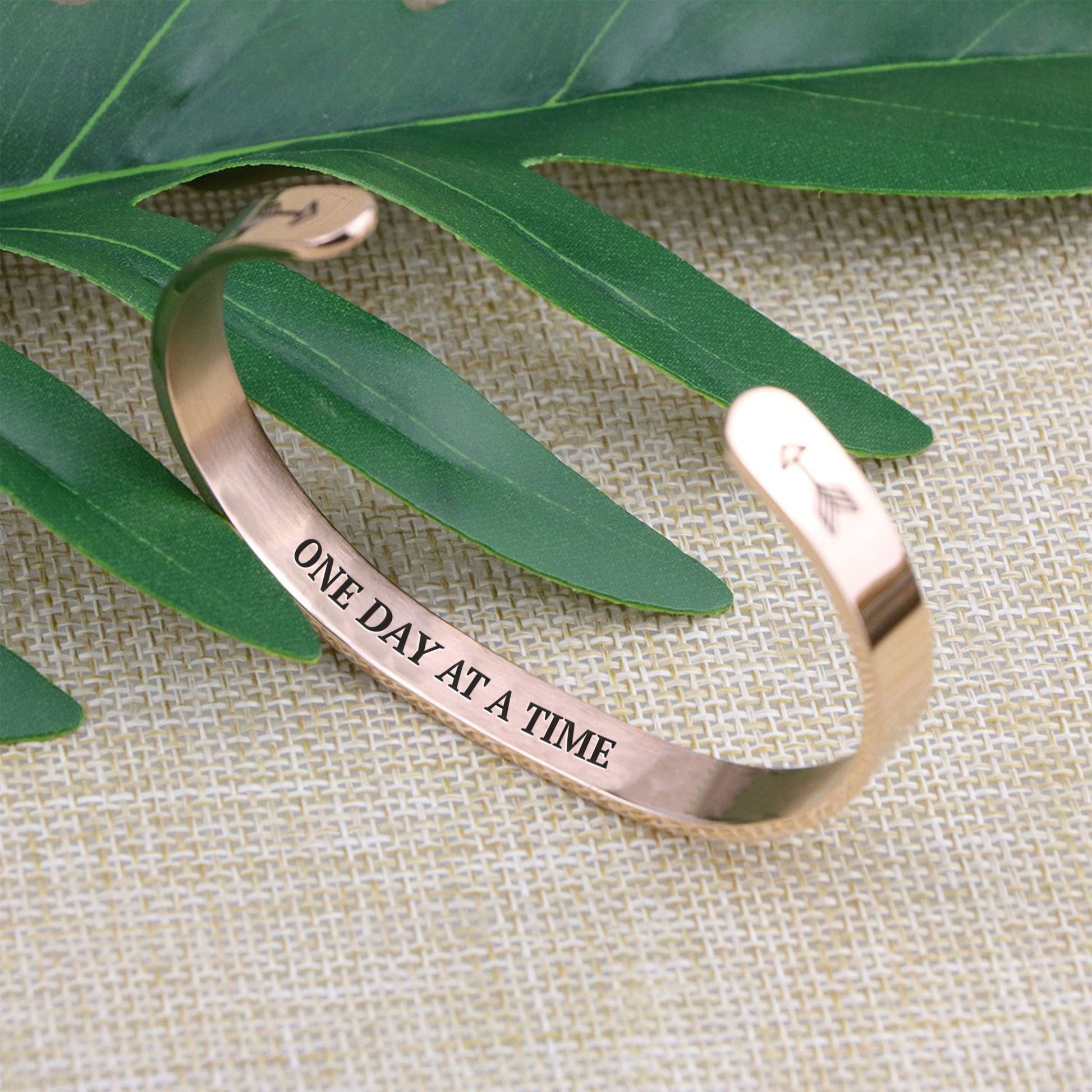 One day at a time bracelet with rose gold plating standing on a burlap surface with a leafy background