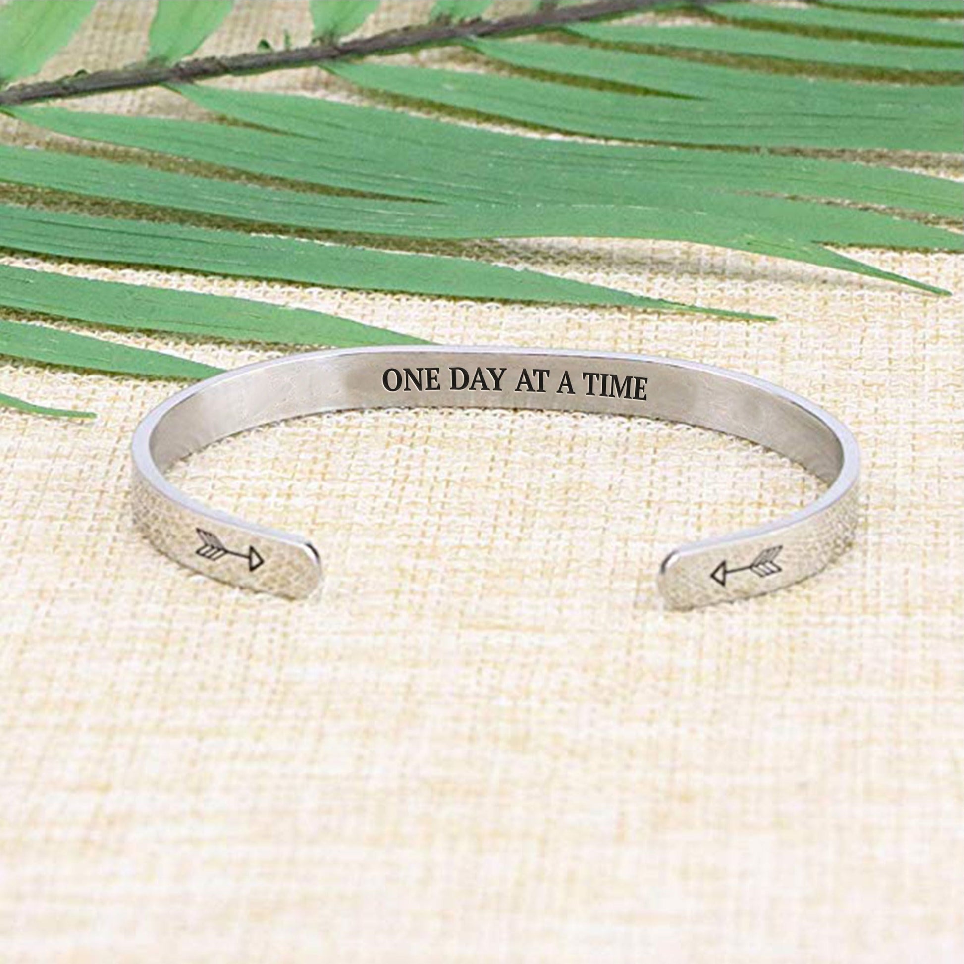 One day at a time bracelet with silver plating laying flat on a burlap surface with a leafy background