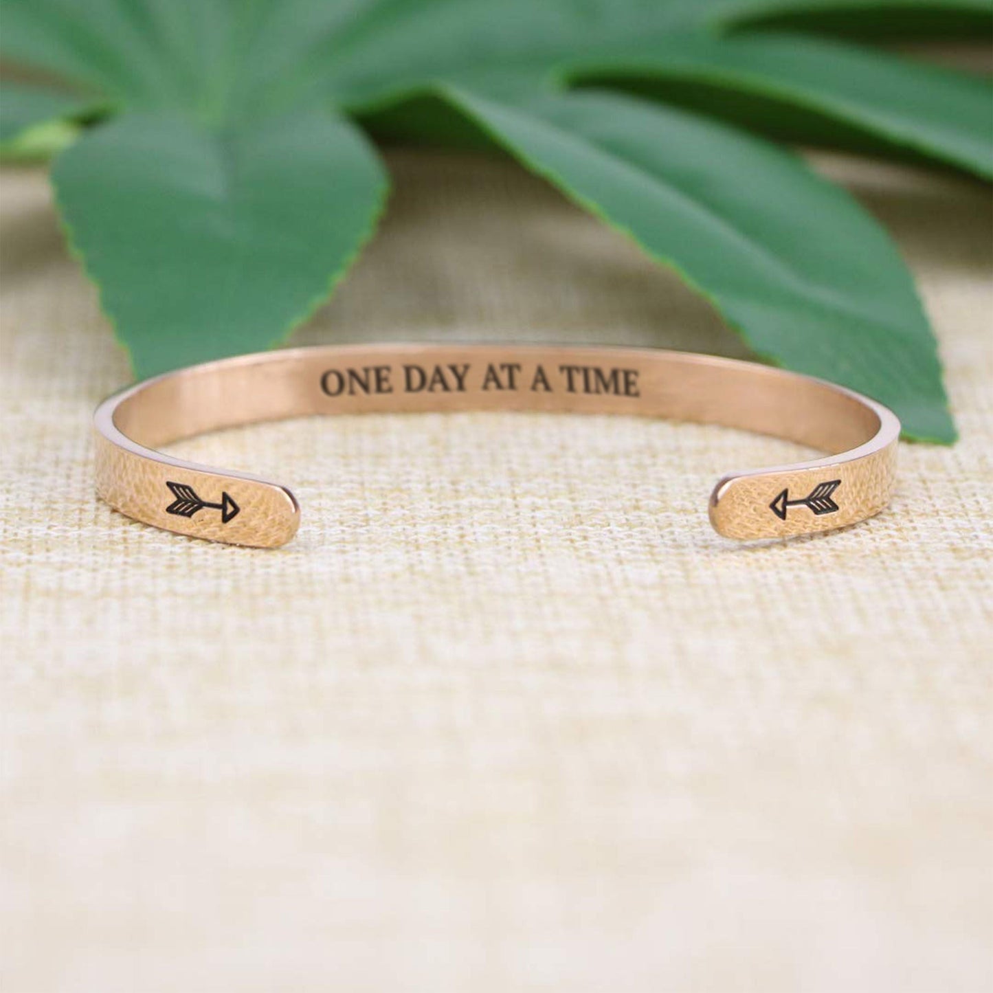 One day at a time bracelet with rose gold plating with arrows in focus on a burlap surface with a leafy background