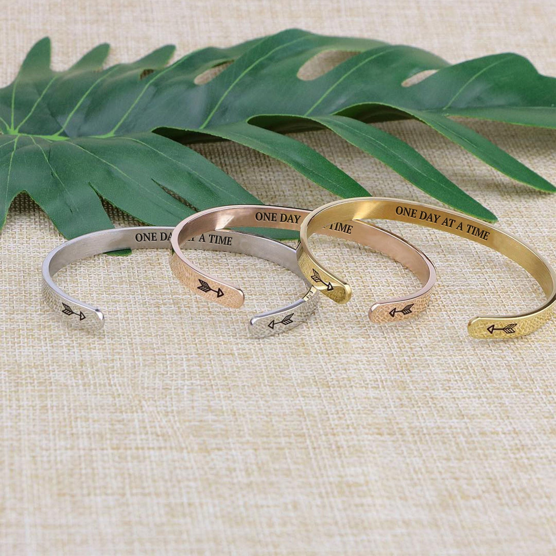 One day at a time bracelets with silver, gold, and rose gold plating staggered on a burlap surface with a leafy background